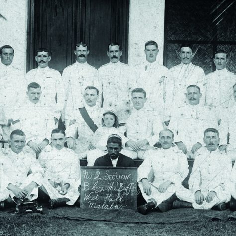 Buffs B Company, Malabar, India. Three rows of men dressed in white. The man centre botton holds a sign, and is dressed in darker colours.